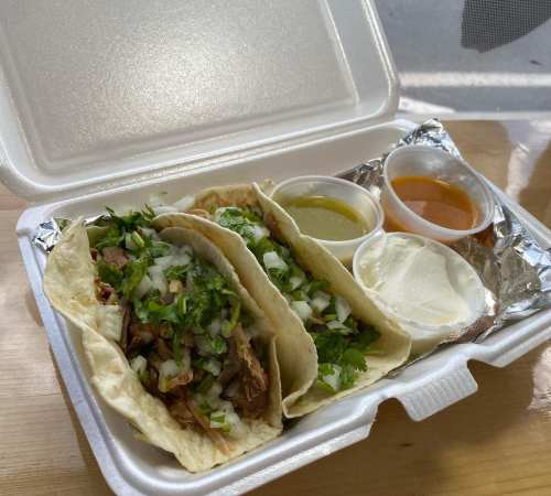 Three tacos filled with meat, topped with onions and cilantro, served with two sauces and a side of sour cream.