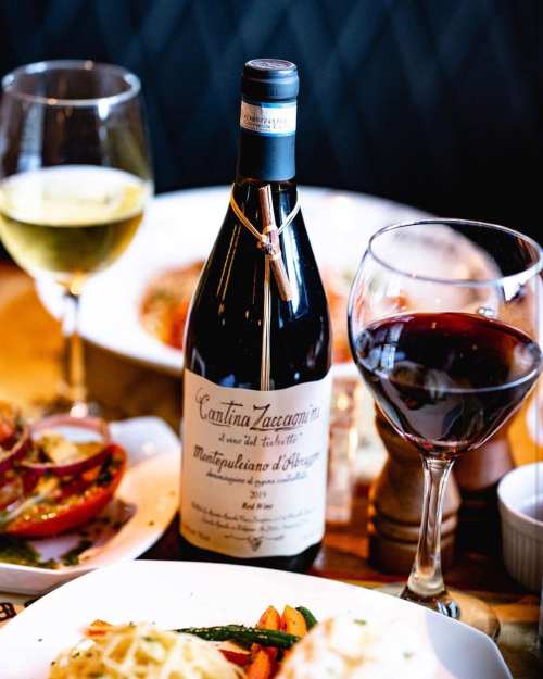 A bottle of red wine with glasses of red and white wine, surrounded by plates of food on a wooden table.