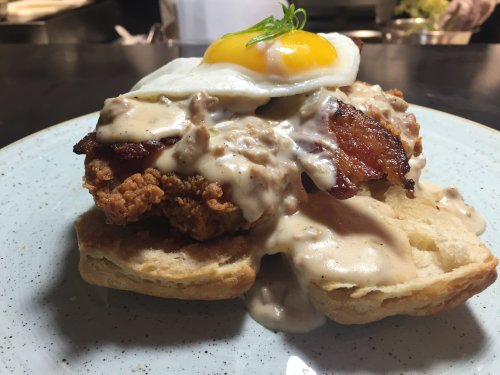A plate of biscuits topped with fried chicken, creamy gravy, crispy bacon, and a sunny-side-up egg.