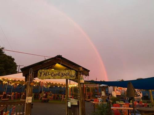 A colorful rainbow arcs over a rustic outdoor dining area with a sign that says "Y'all Come Back Now."