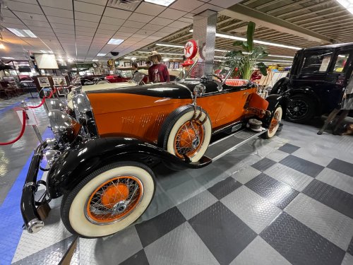 A vintage orange and black convertible car displayed in a classic car museum with checkered flooring.