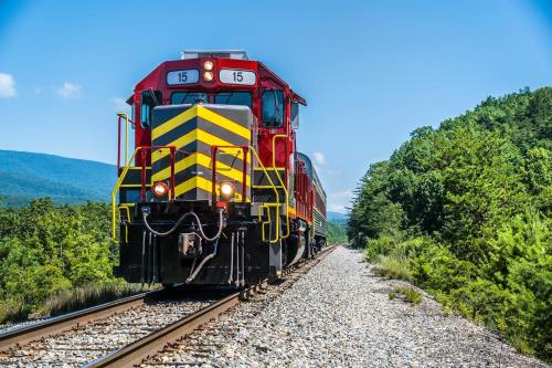 A red and yellow train on tracks, surrounded by greenery and mountains under a clear blue sky.