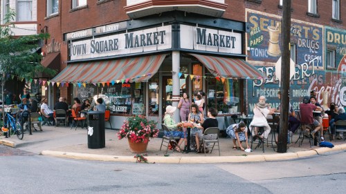 A bustling market with outdoor seating, colorful decorations, and people enjoying food and drinks on a sunny day.