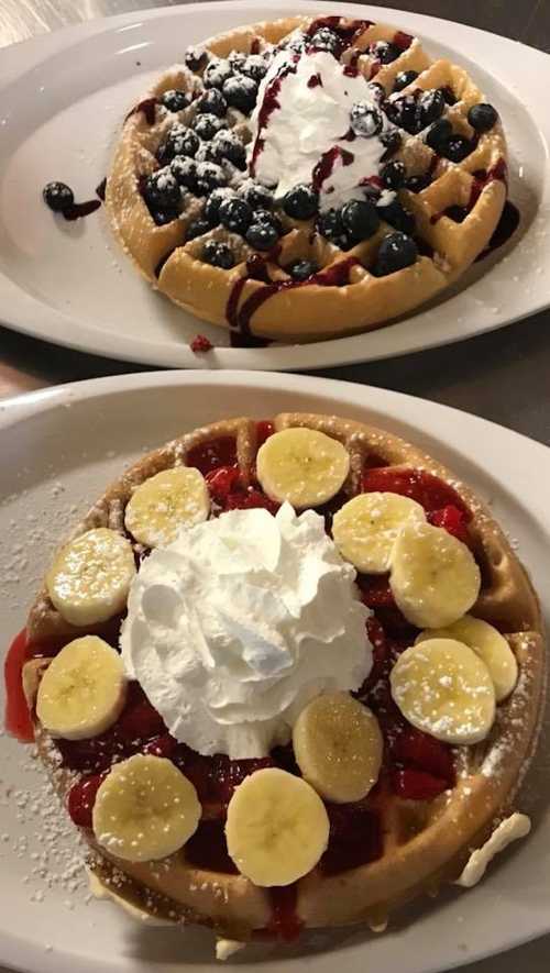 Two plates of waffles: one topped with blueberries and whipped cream, the other with bananas and strawberries.