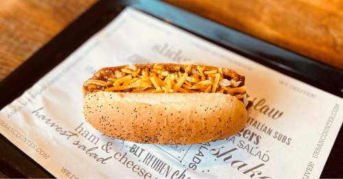 A poppy seed hot dog bun filled with chili and topped with shredded cheese, served on a printed paper tray.