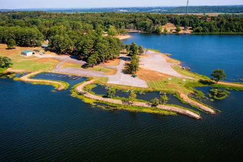 Aerial view of a serene lake with a boat ramp, grassy areas, and wooded surroundings on a sunny day.
