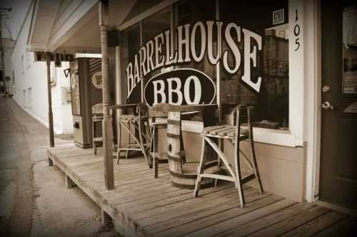 A rustic BBQ restaurant with wooden seating and barrels on a porch, featuring a large sign that reads "Barrelhouse BBQ."