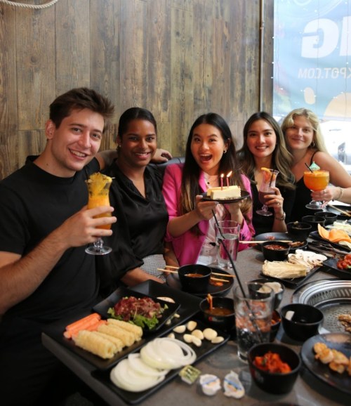 A group of five friends celebrating with drinks and a cake at a restaurant, surrounded by various dishes.