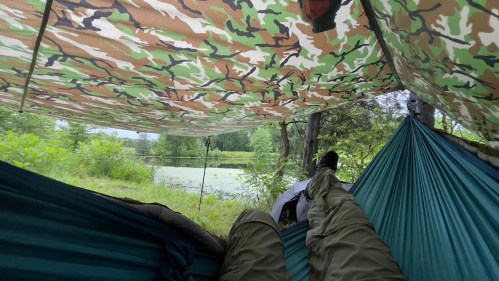 A person relaxes in a hammock under a camouflage tarp, surrounded by greenery and a calm pond in the background.
