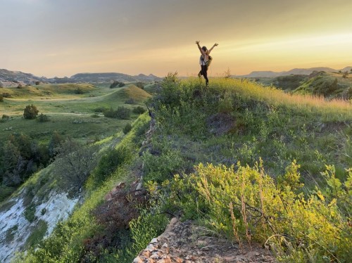 A person stands on a grassy hilltop at sunset, arms raised, with rolling hills and a colorful sky in the background.