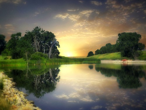 A serene river at sunset, reflecting trees and clouds, with a rocky shore and lush greenery in the background.