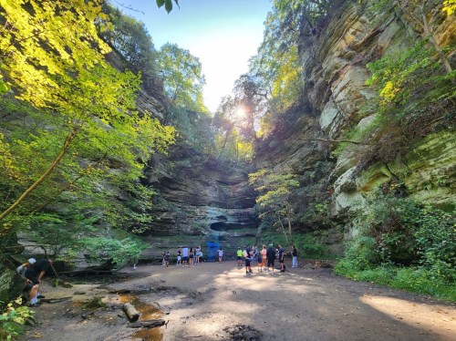 A group of people gathers at a scenic canyon with lush greenery and sunlight filtering through the trees.
