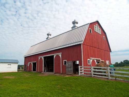 A large red barn with a metal roof, surrounded by green grass and a white fence under a cloudy sky.