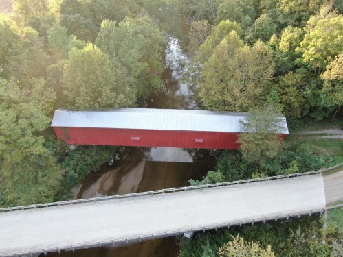 Aerial view of a red covered bridge over a calm river, surrounded by lush green trees and a nearby road.