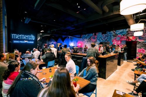 A lively lounge scene with people socializing, a bar, and vibrant floral murals in the background.