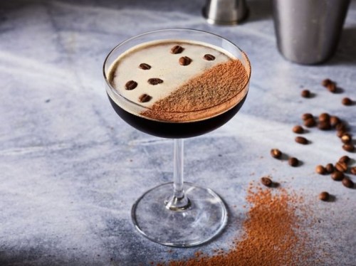 A cocktail in a coupe glass topped with coffee beans and cocoa powder, set on a marble surface with scattered coffee beans.