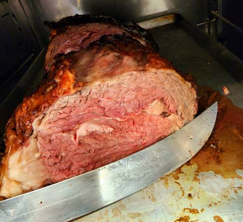 A large, cooked roast beef with a shiny, browned exterior, partially sliced on a metal tray.