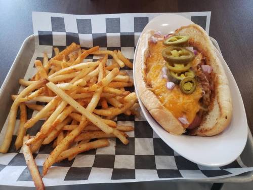 A loaded hot dog topped with cheese, jalapeños, and onions, served with a side of seasoned French fries on a checkered tray.