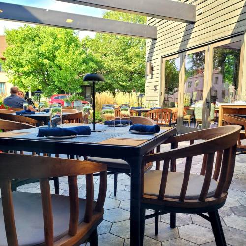 Outdoor dining area with wooden tables and chairs, surrounded by greenery and a sunny atmosphere.