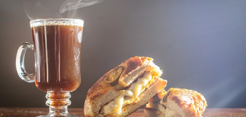 A steaming cup of coffee beside a toasted sandwich filled with cheese and pickles, set against a soft-lit background.