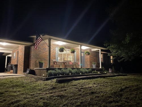 A well-lit brick house at night, featuring a porch with seating, American flag, and landscaped garden.