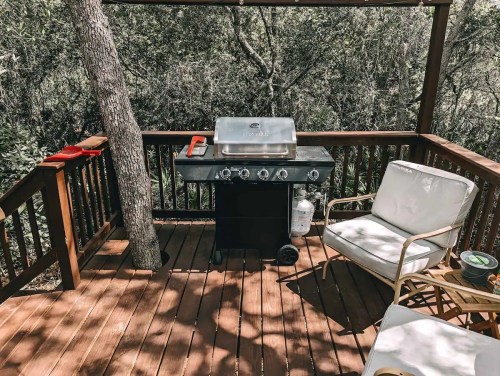 A wooden deck with a grill, two chairs, and a tree, surrounded by greenery.