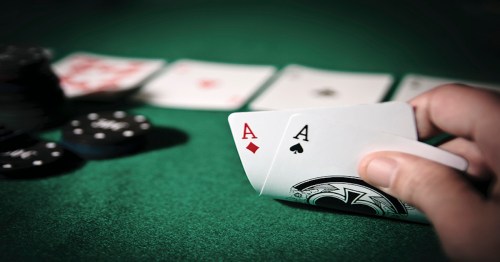 A hand holding two aces over a green felt poker table with playing cards and chips in the background.