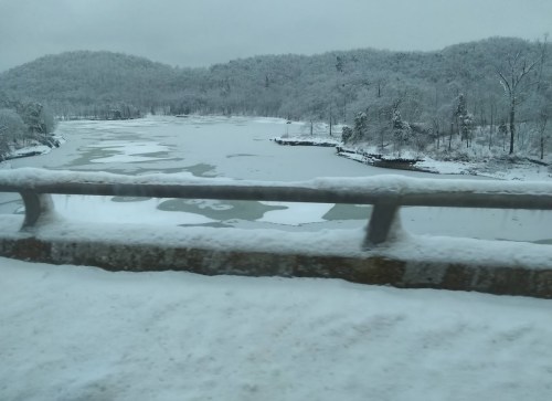 Snow-covered landscape with a frozen river and hills in the background, viewed from a bridge.
