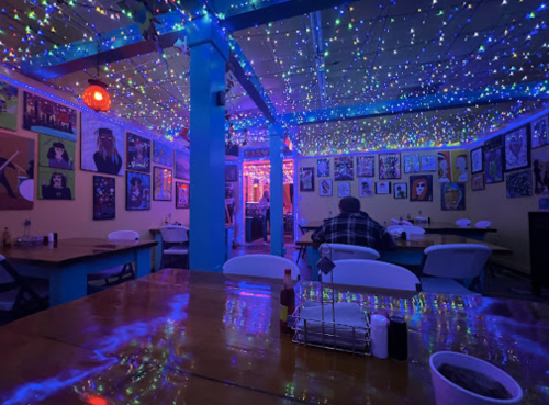 A colorful restaurant interior with string lights, vibrant artwork on the walls, and empty tables.
