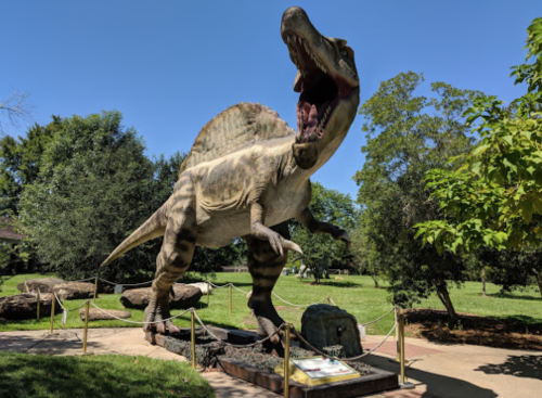 A large dinosaur statue with a raised head and open mouth, set in a green park with trees and rocks.