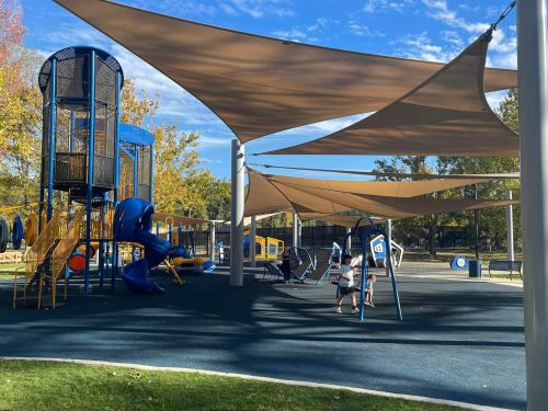 A playground with colorful equipment and shade sails, featuring slides, swings, and children playing under a blue sky.