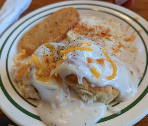 A plate of biscuits smothered in creamy gravy, topped with shredded cheese, alongside a hash brown and eggs.