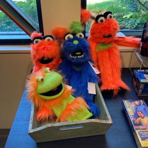A collection of colorful puppet monsters in a wooden tray, featuring bright orange, green, and blue fur.