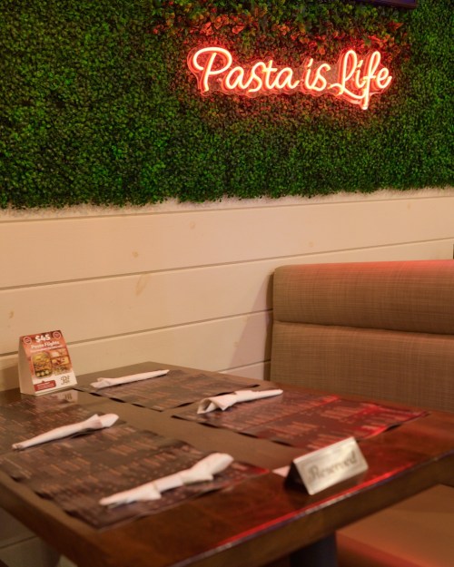 A cozy restaurant table set for two, with a neon sign reading "Pasta is Life" on a green wall.