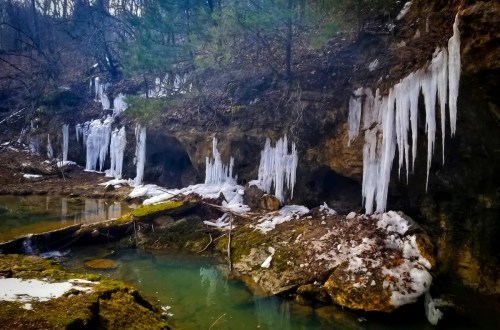 Icicles hang from rocky cliffs beside a serene, icy blue stream in a winter landscape.