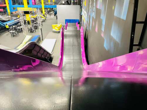 A colorful indoor slide descends into a vibrant play area with seating and activity spaces visible in the background.