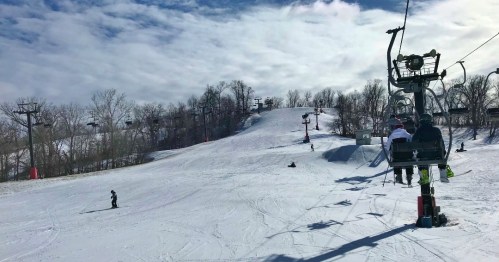 A snowy ski slope with a chairlift, skiers, and trees under a partly cloudy sky.