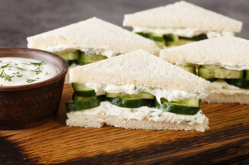 Three cucumber sandwiches with creamy filling, served on a wooden board alongside a small bowl of dip.