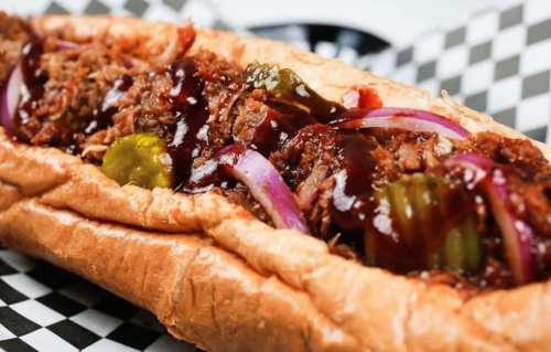 A close-up of a loaded sandwich with pulled meat, barbecue sauce, pickles, and red onions in a toasted bun.