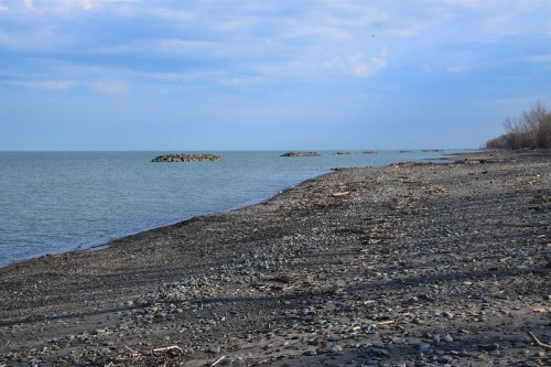 A rocky shoreline extends along a calm lake under a cloudy sky, with scattered debris and distant rocky formations.