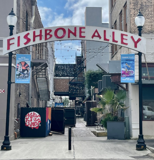 Archway sign reading "Fishbone Alley" with string lights and colorful posters in a narrow alleyway.