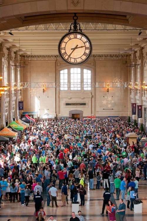 A large crowd gathers in a spacious hall with a clock, colorful tents, and bright lighting.