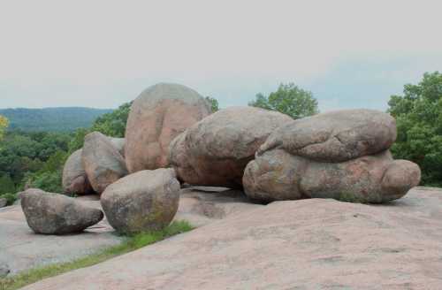 A cluster of large, smooth boulders on a rocky surface, surrounded by greenery and distant hills under a cloudy sky.