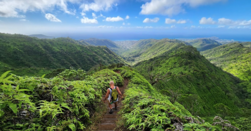 A hiker descends a lush, green mountain trail with a panoramic view of valleys and ocean in the background.