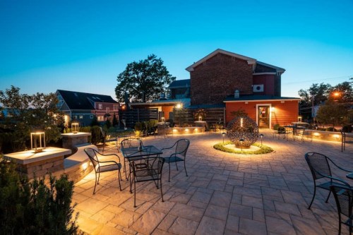 A serene outdoor patio at dusk, featuring stone pathways, seating, and decorative lighting around a central sculpture.