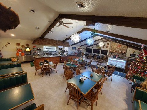 A cozy restaurant interior with green tables, wooden chairs, a festive Christmas tree, and a counter in the background.
