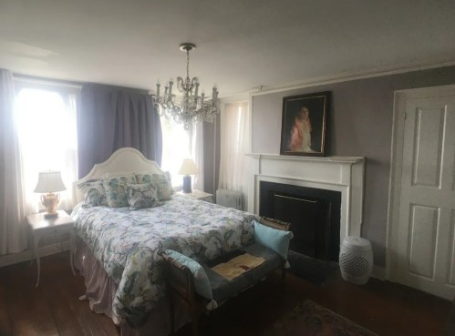 Cozy bedroom featuring a floral bedspread, chandelier, and a fireplace with a painting above it. Natural light from windows.