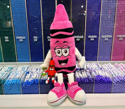 A large pink crayon plush toy with a smiling face sits next to a smaller red crayon plush on a colorful background of crayons.