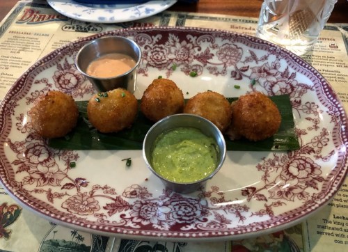 A decorative plate with five golden fried balls, served with two dipping sauces, on a bed of green leaves.