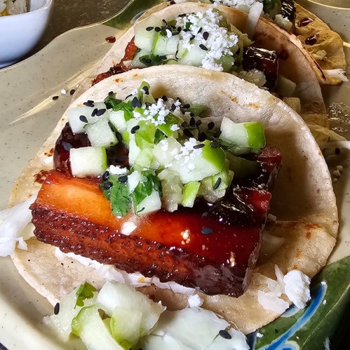 Three tacos filled with pork, topped with diced cucumber, cilantro, and sesame seeds on a plate.
